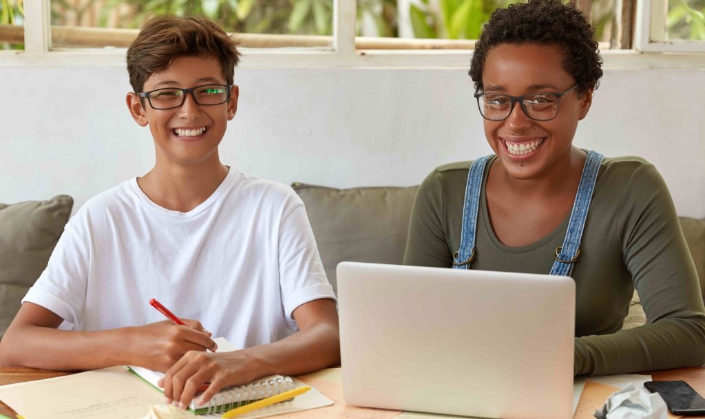 Mixed race students of high school learn together at coworking space, watch training webinar on laptop computer, write records in spiral notebook, find creative solution, have toothy smiles. high school homeschool curriculum