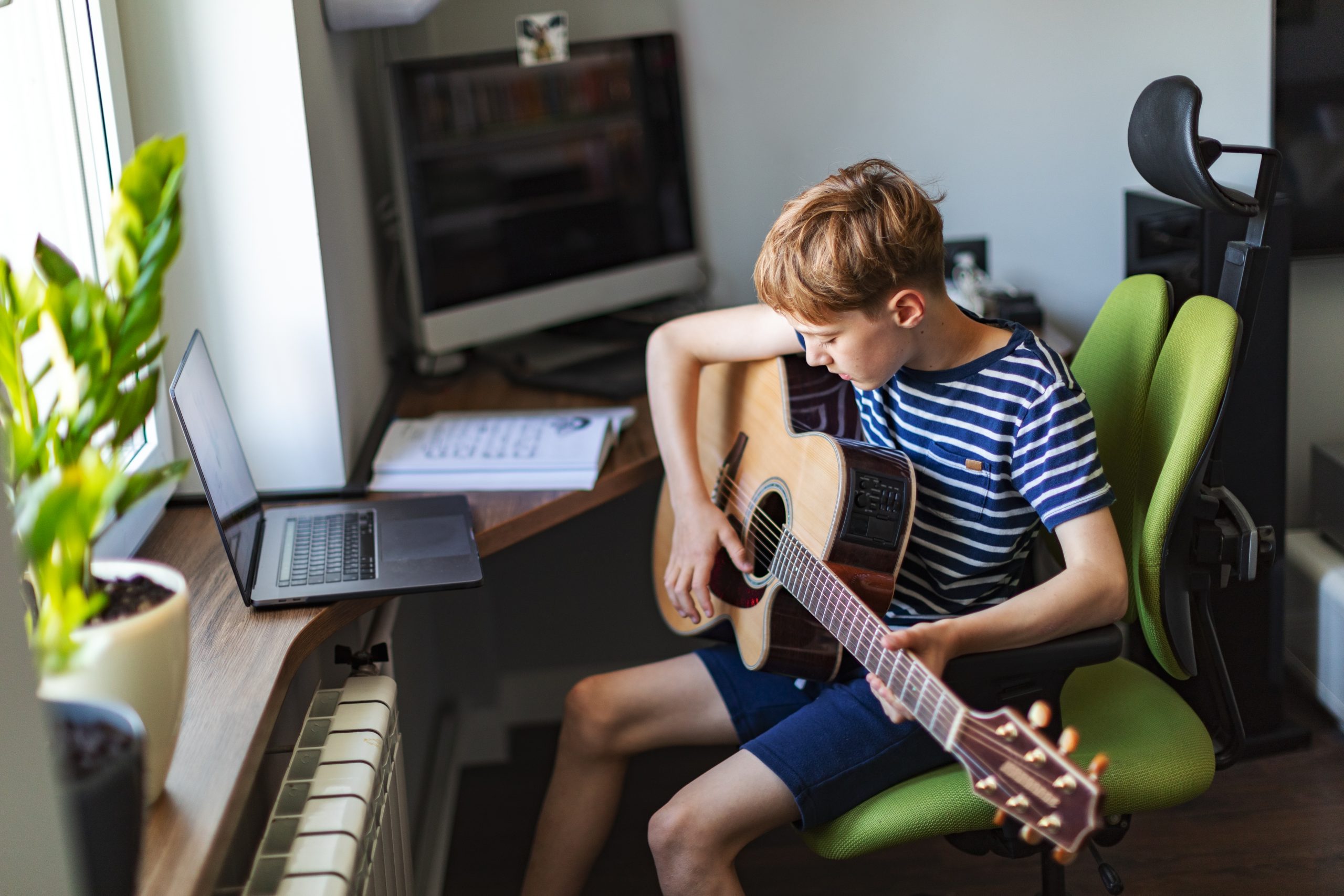 Elementary school age boy playing guitar at home using laptop